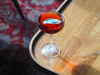 An elegant red-coloured cocktail in a stemmed glass on the corner of a wooden table, with patterned carpet in the background