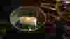 Golden liquid being poured from a glass jug into a bowl of beautiful food against a dark background