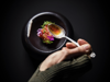 Overhead shot of a bowl of beautiful, colourful food against a very dark background, with a hand holding a spoon