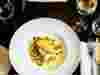 Overhead shot of wide-rimmed white bowl on wooden table top, with pan-fried white fish and mussels in a creamy sauce, accompanied by glass of white wine, linen napkin and cutlery