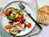 Overhead shot of aubergine, tomatoes, red onion and basil on plain white plate sitting on folded pale blue cloth napkin. Two lightly toasted slices of bread sit beside the plate.