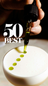 Close-up of a hand adding droplets of green oil onto the frothy top of a cocktail with 50 Best logo