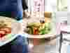 Person in apron holding two plates of salad with table in background