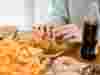 Person eating plate of fried chicken and chips, with other unhealthy food on table